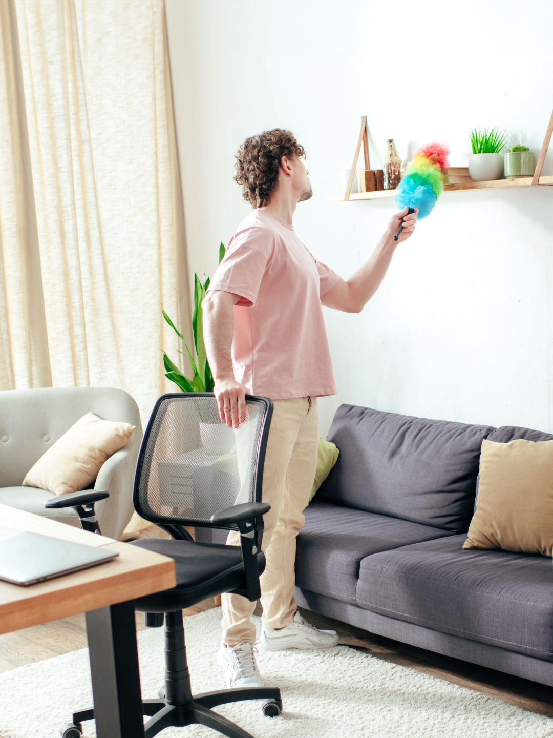 A person standing in a bright, modern living room is using a colorful duster to clean a shelf. The room features a gray sofa, a beige chair, a wooden table with a laptop, and decorative plants on the shelf. Natural light is coming through the curtains.