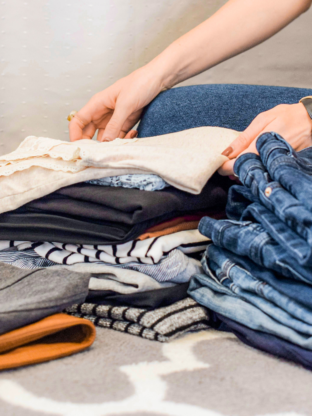 A person arranging a stack of neatly folded clothes, including various tops and denim, on a patterned rug.