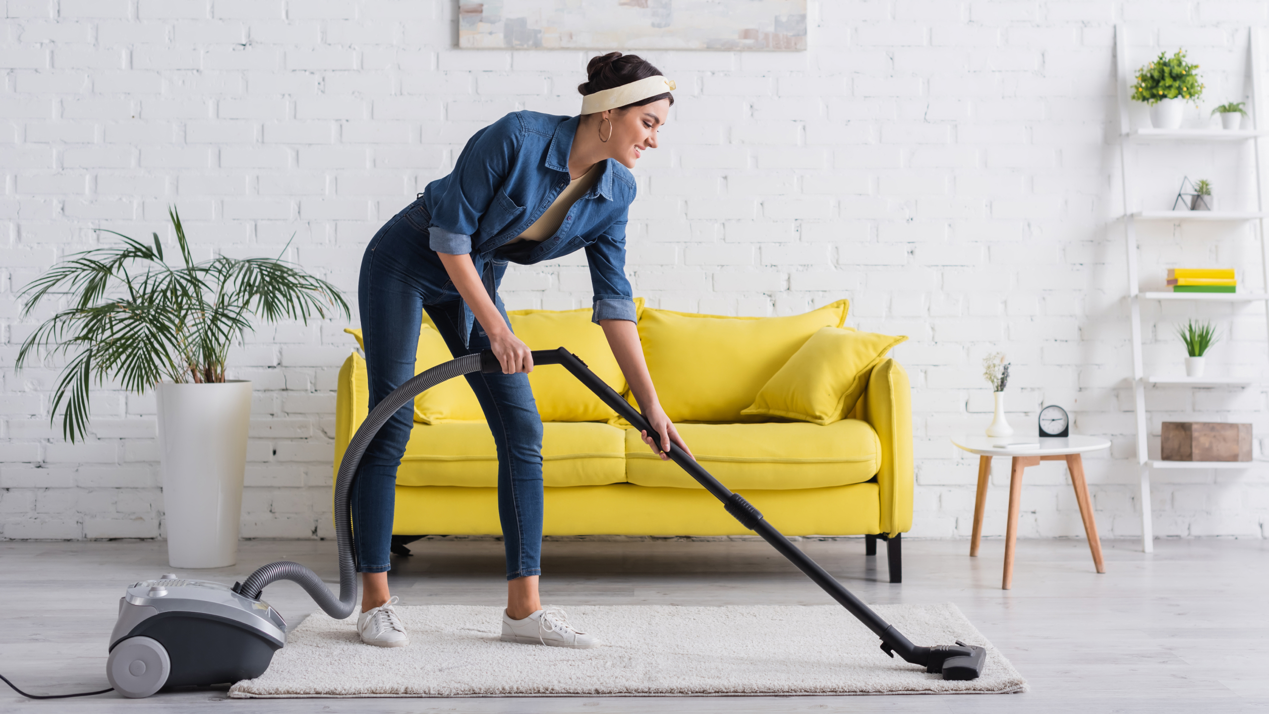 A woman in a denim outfit vacuuming a light-colored carpet in a bright living room with a yellow sofa and a potted plant.